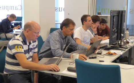 A group of men sitting at a desk looking at laptops and monitors