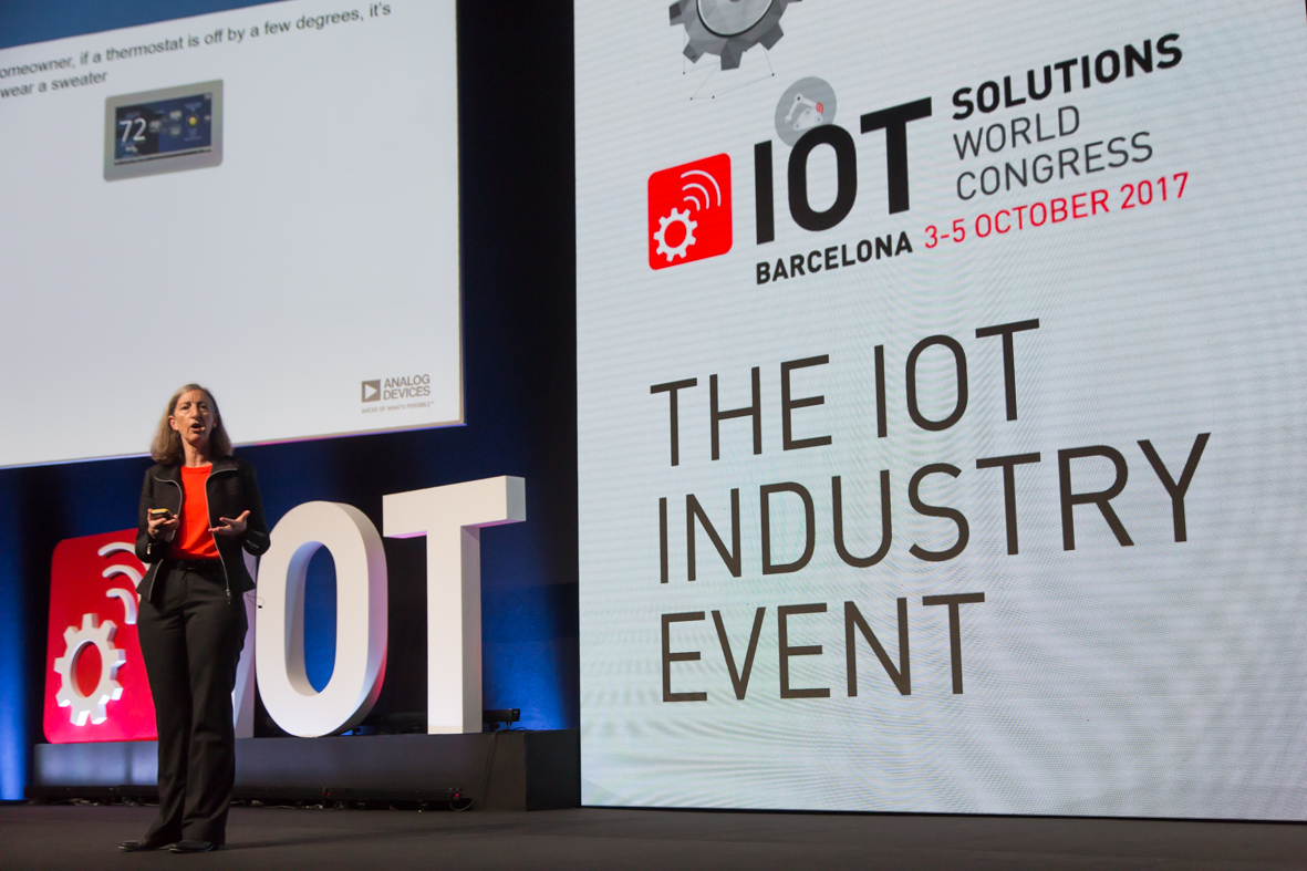 Woman speaking on a stage with "The IoT Industry Event" on a banner behind her.