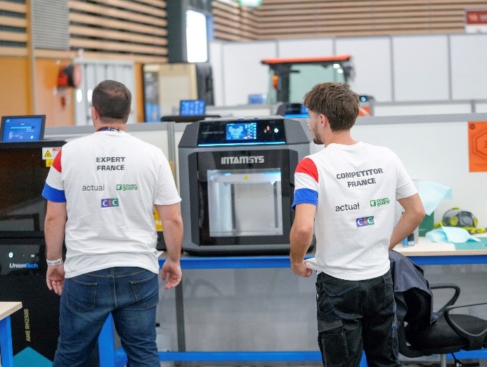 Two additive manufacturing technicians stand at additive manufacturing machines.