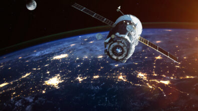 A spacecraft with solar panels extended orbiting above an Earth lit by city lights.