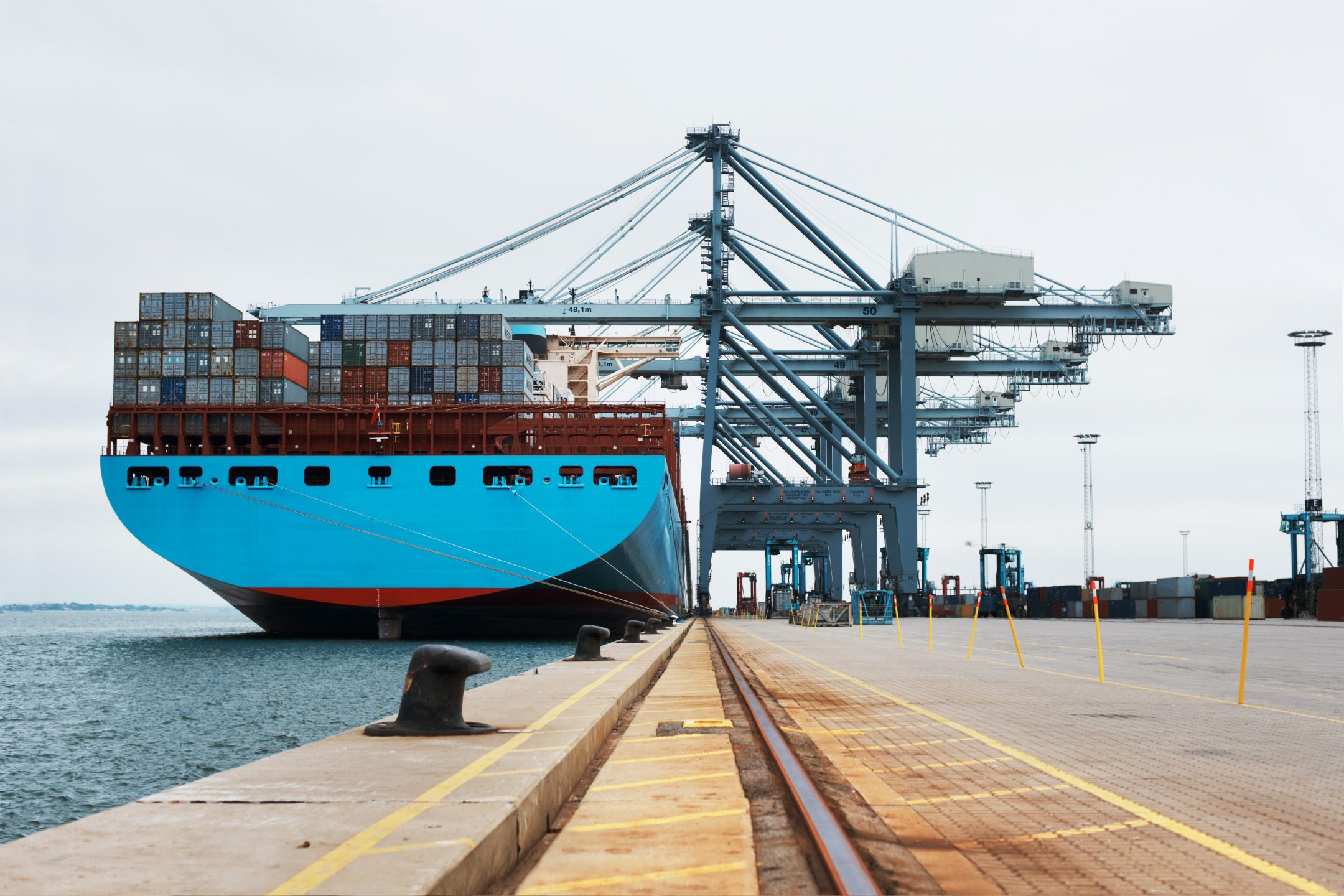 Blue container ship docked at a harbor being serviced by cranes.