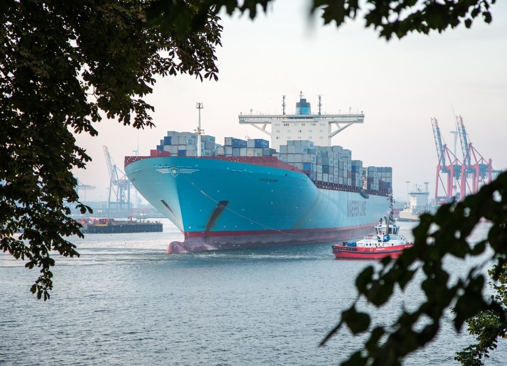 A light blue container ship sailing into a harbor alongside a tug.