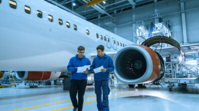 Two aircraft mechanics with tablets talking with each other aside an airplane in a hangar.