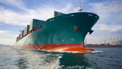 The bow-view of a blue-green cargo ship sailing out of a port.