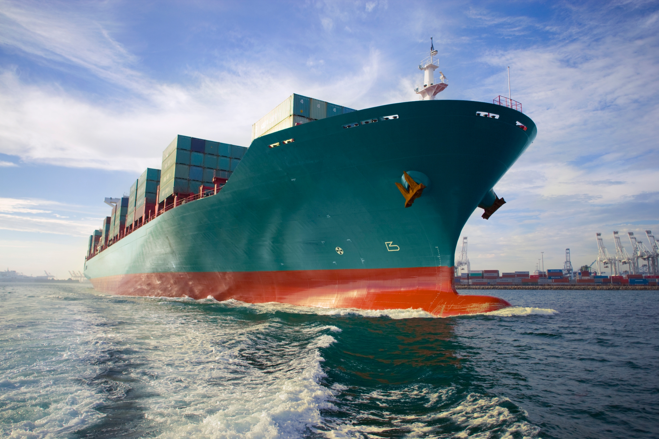 The bow-view of a blue-green cargo ship sailing out of a port.