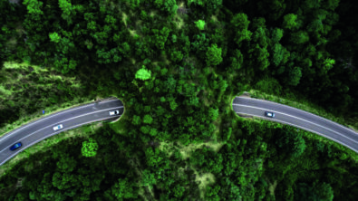 A bird's eye view of a road curving through a green forest with a few cars driving along it.