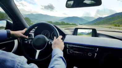 A car dashboard with a person behind the steering wheel, driving through a hilly landscape.