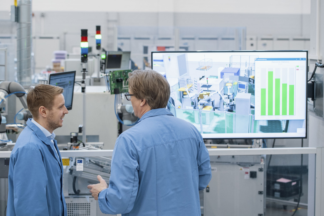 Two Siemens Electronics Factory Erlangen workers discussing the digital twin of a factory floor on a nearby screen.