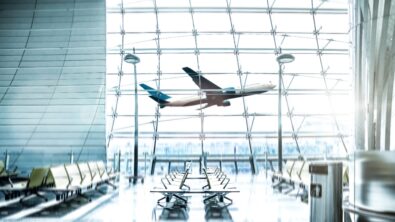 An airport waiting lounge with an airplane flying behind a large window in the daytime.