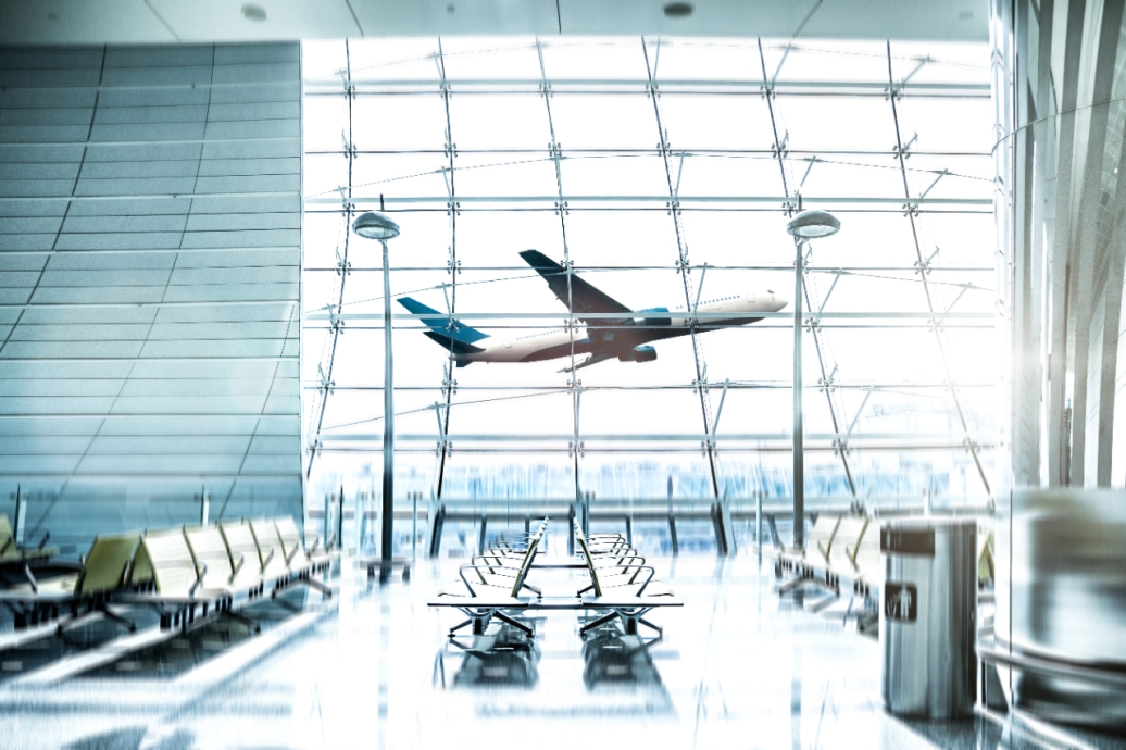 An airport waiting lounge with an airplane flying behind a large window in the daytime.