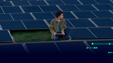 A person crouching in the middle of an array of solar panels, holding an electronic pad in their hands.