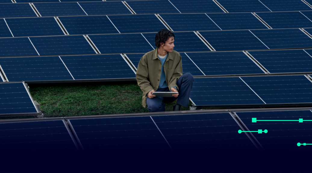A person crouching in the middle of an array of solar panels, holding an electronic pad in their hands.