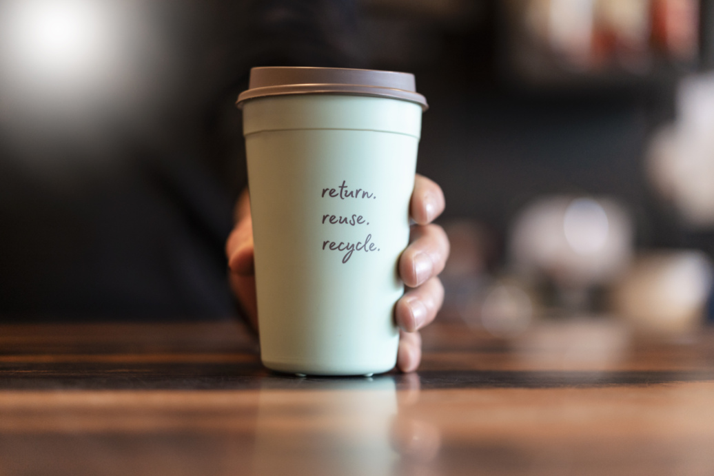 Close-up of a hand holding a cup that says "Return, reuse, recycle."