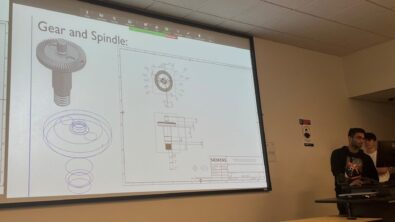 Student presenting a slide of a gear and spindle design in NX, with both the 3D model and the 2D sketch lines.