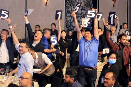 A large group of people hold up cards with various single-digit numbers on them while in a banquet room at AP Converge.