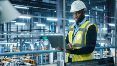 AI in manufacturing is represented by a man in a factory holding a laptop to monitor automobile production.