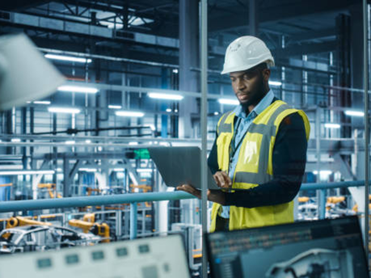 AI in manufacturing is represented by a man in a factory holding a laptop to monitor automobile production.