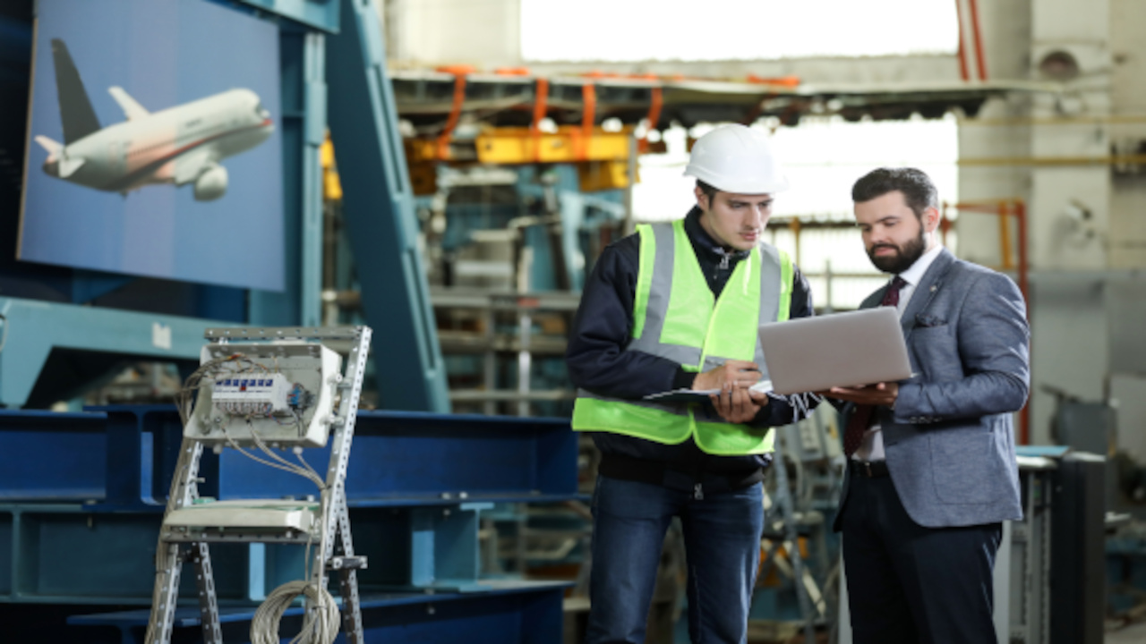 Two workers, one in a reflective vest and a hard hat and the other in a suit holding a laptop, confer about aerospace manufacturing