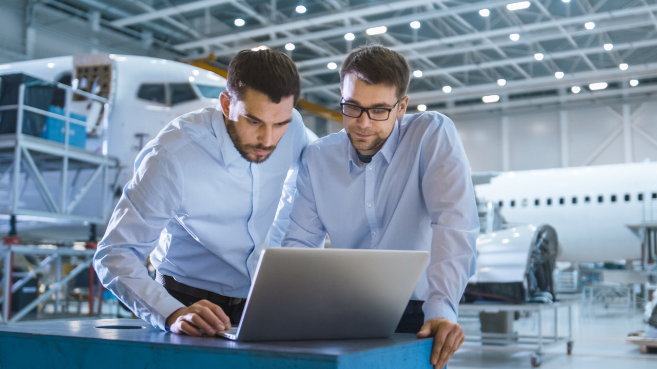 Two workers in an aircraft hangar look at a laptop