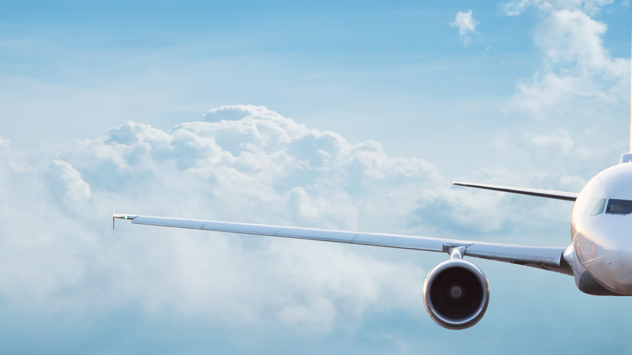 A commercial airplane is flying directly toward the camer with a light blue sky and clouds in the background.