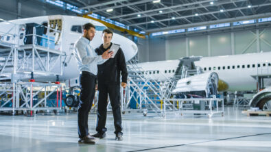 Two aerospace engineers reviewing a digital plan on a tablet inside an aircraft manufacturing facility—illustrating collaboration and model-based production planning.