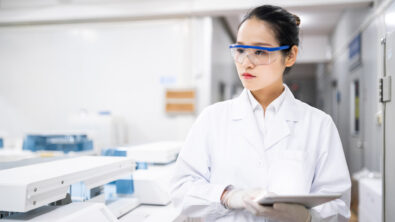 Woman wearing a lab coat and protective eye wear walking through a lab focusing on quality management.