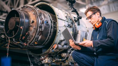 An aircraft engineering working on a laptop aside an unfinished jet engine.