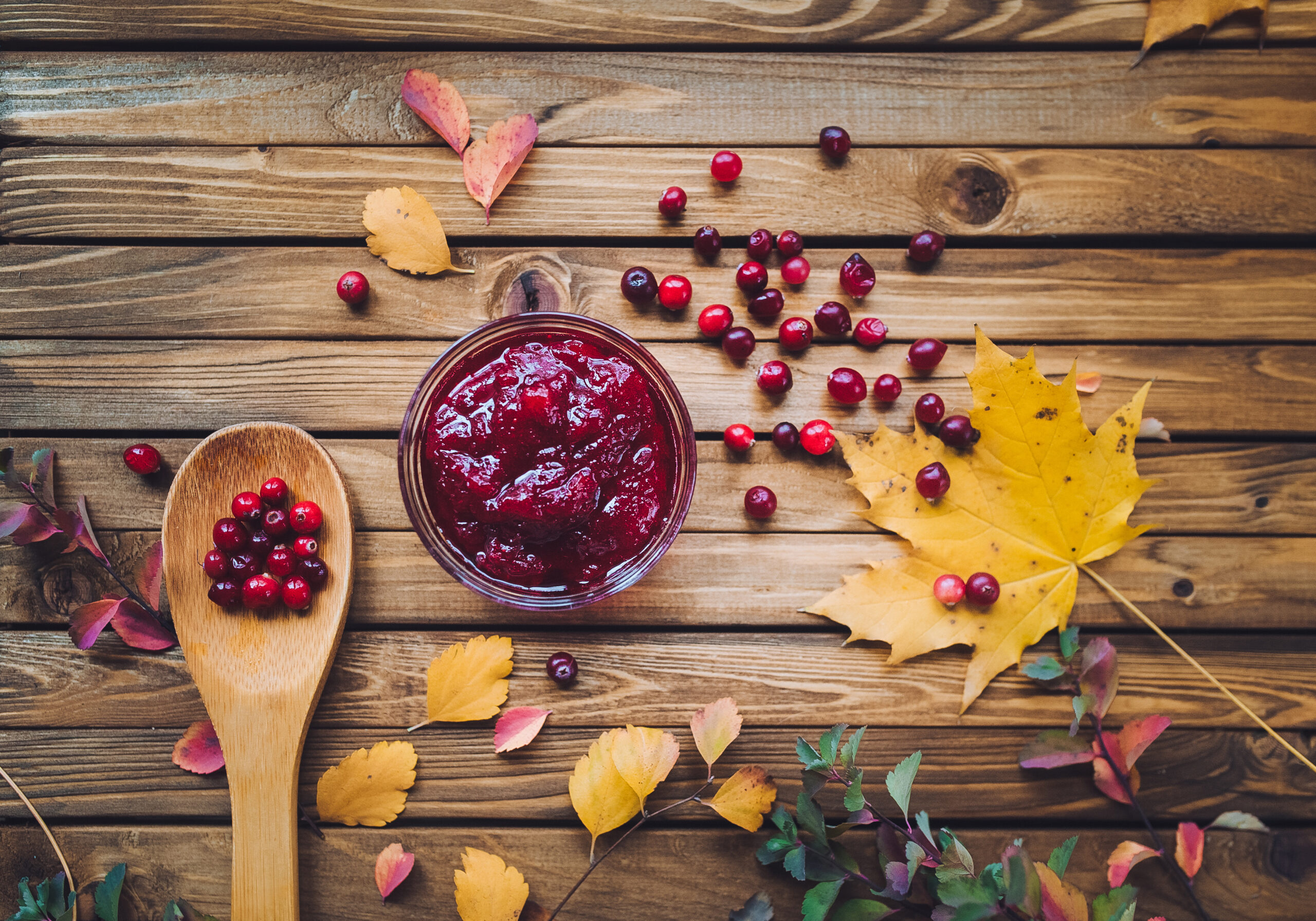 A wood-plank table with a wooden spoon holding several cranberries beside a small glass bowl holding prepared cranberry sauce surrounded by loose cranberries and a small assortment of leaves in autumn colors