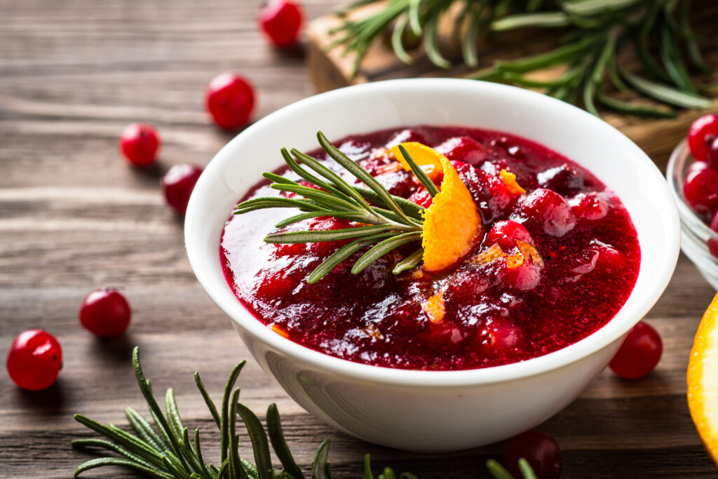 A bowl of homemade cranberry sauce with a sprig of rosemary and orange peel garnish  in a white ceramic bowl on a wooden table with scattered loose cranberries and rosemary