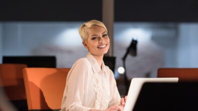Business woman in a dark office sitting at her desk smiling in front of a laptop