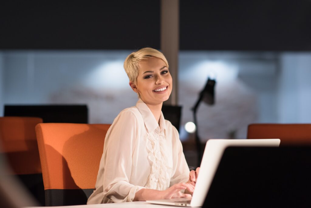Business woman in a dark office sitting at her desk smiling in front of a laptop