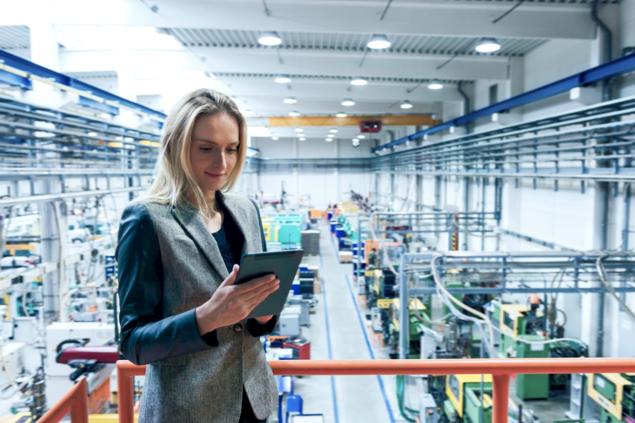 Business woman in manufacturing plant using a tablet, standing in front of heavy machinery
