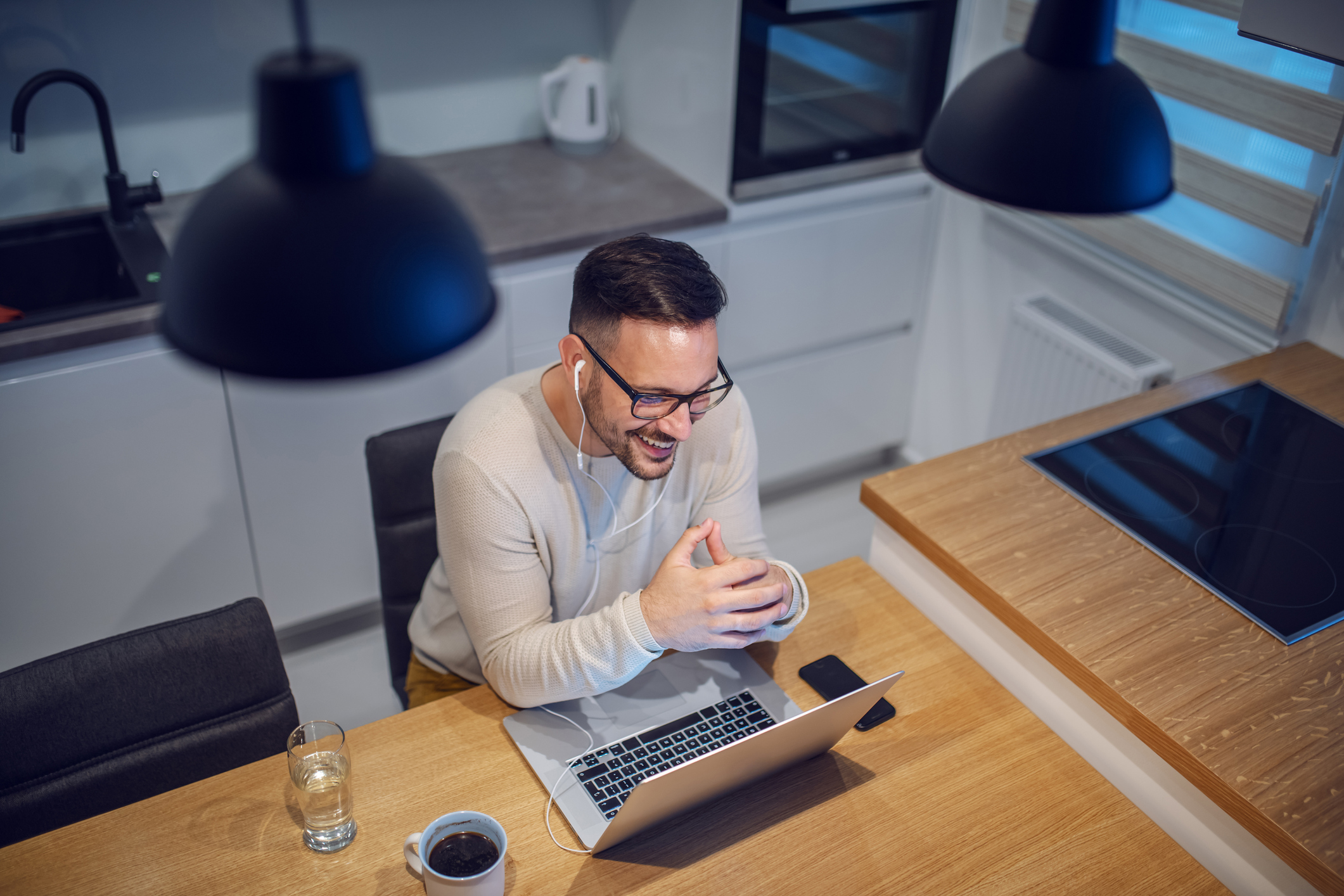 Top view of attractive smiling caucasian man sitting at dining table in kitchen and having video call with his girlfriend. On table are laptop, glass with water and mug with coffee.