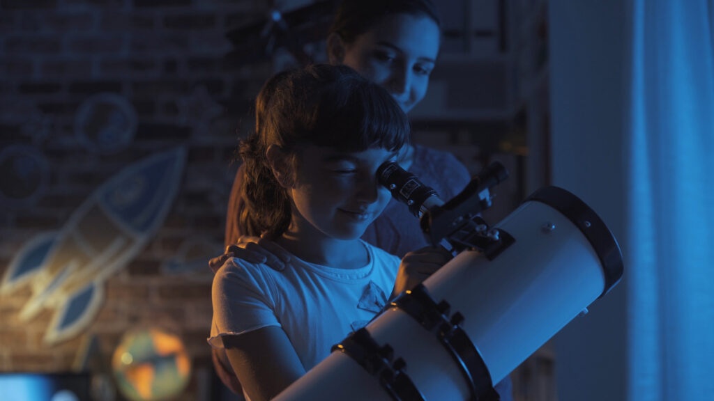 A young child sits by a window at night, peering through a telescope aimed at a star-filled sky. The room is softly lit, with books and astronomy posters nearby, capturing the wonder and curiosity of early stargazing.