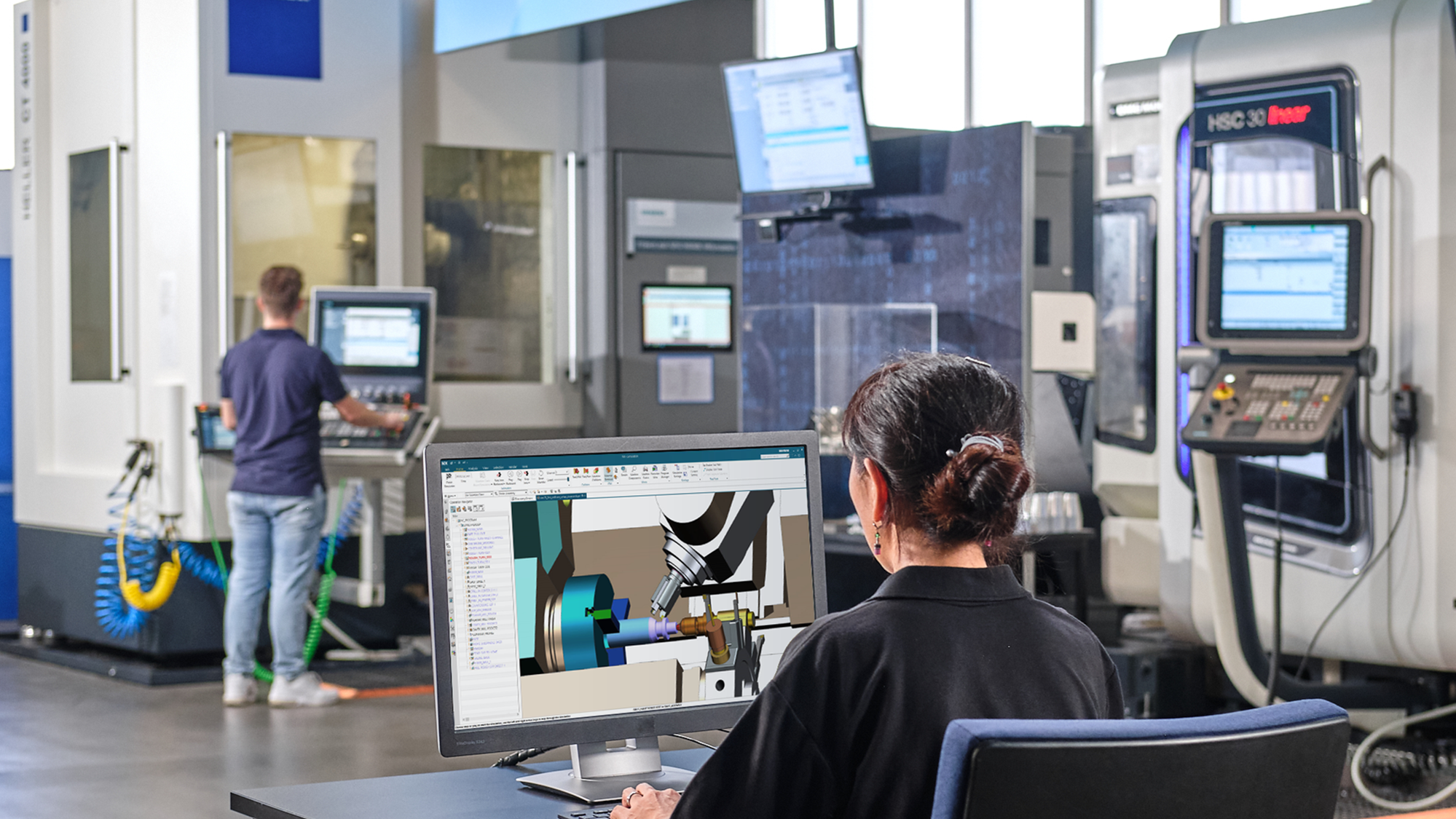 Two engineers in a CNC machining workshop: a woman reviewing a CAM software model on a monitor and a man operating a CNC machine control panel