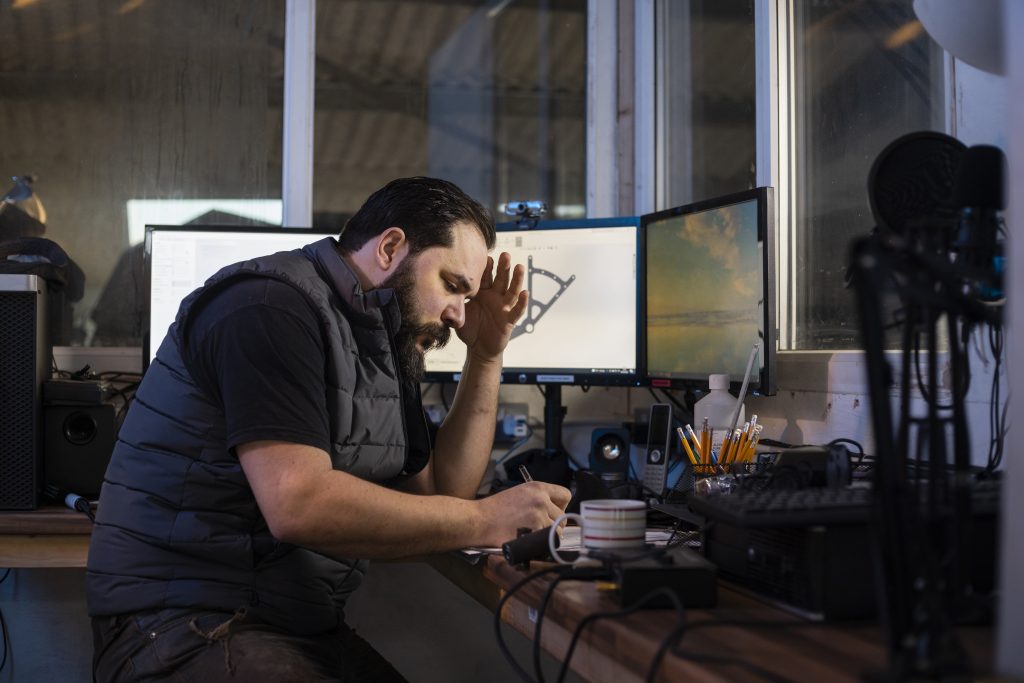 A frustrated engineer sits at a dual-monitor workstation displaying CAD simulation data, illustrating the complexity of traditional iterative engineering design workflows.