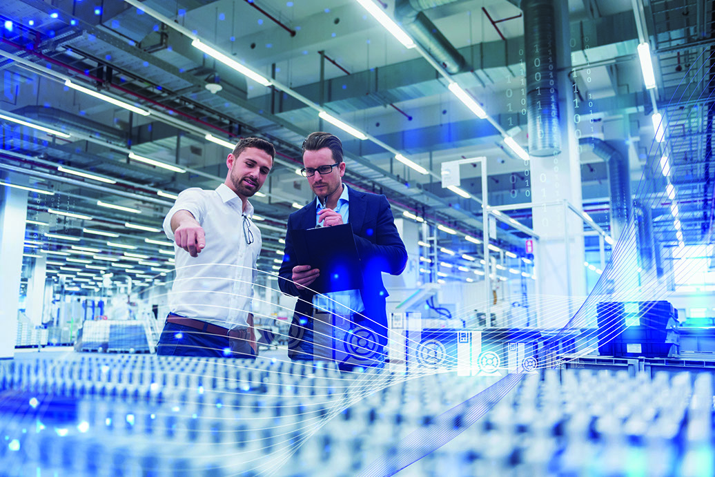 Two business men standing in a factory. One is pointing and the other takes notes on a tablet.