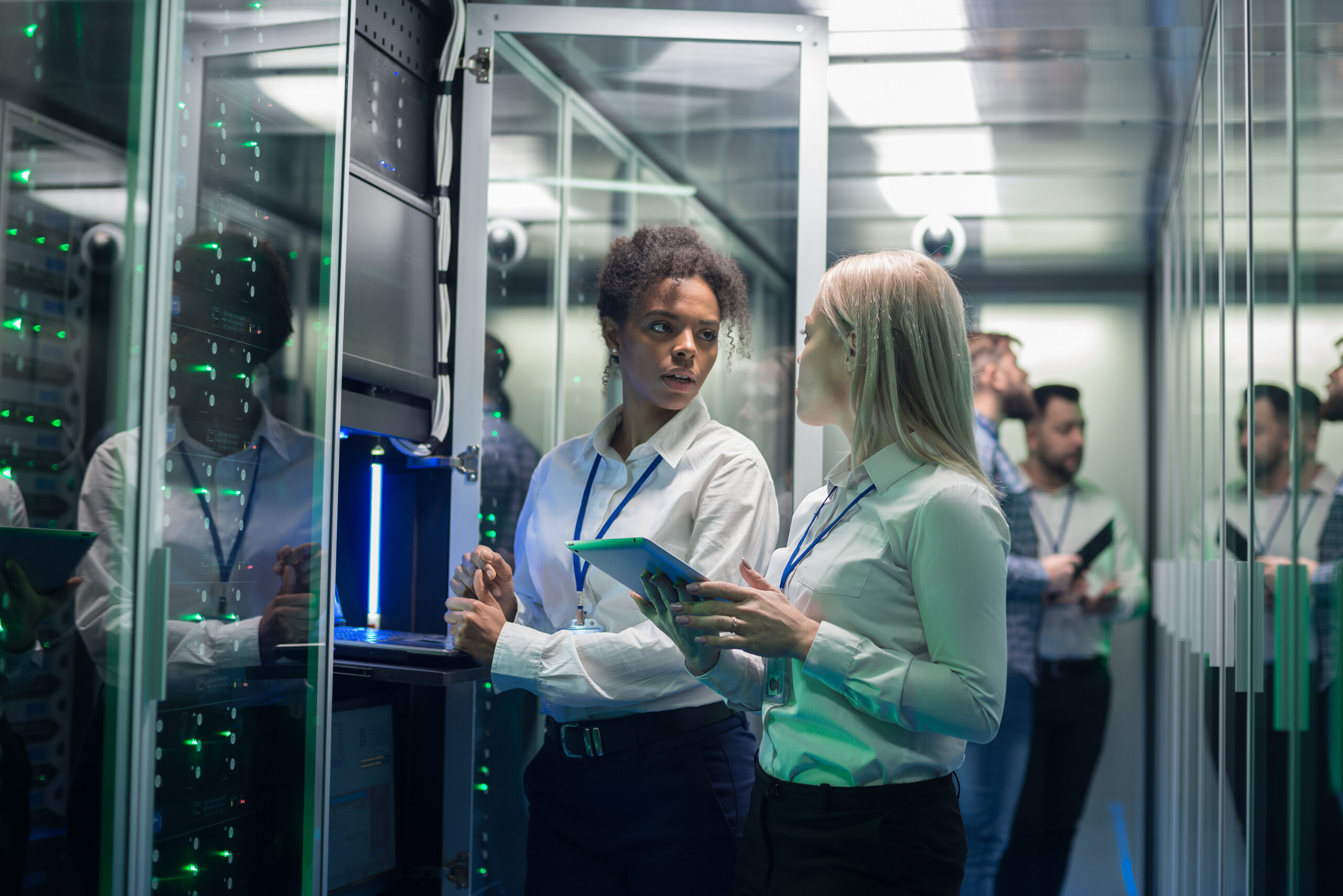 Medium shot of two women working in a data center with rows of server racks and checking the equipment and discussing their work