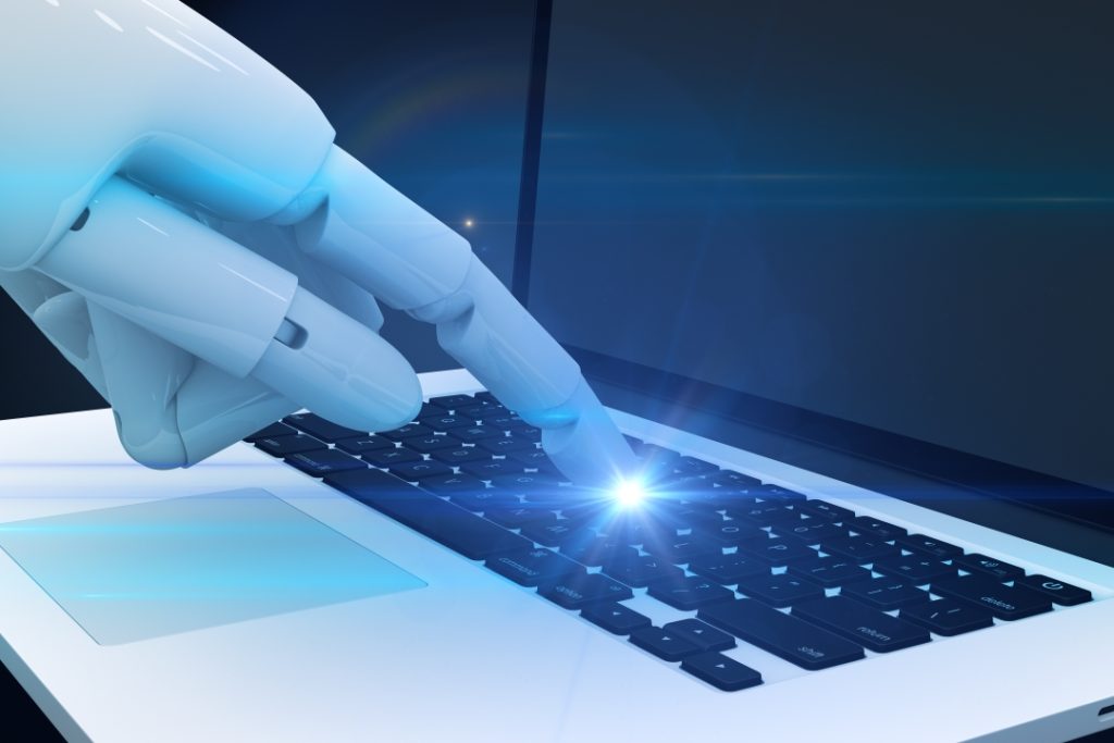 A translucent robotic hand with a glowing blue finger pointing at a laptop keyboard. The laptop features a white body with a dark keyboard illuminated by a bright blue light emanating from the robot's fingertip. The background is dark blue, creating a tech-forward atmosphere that suggests artificial intelligence, automation, or digital interaction between humans and machines.