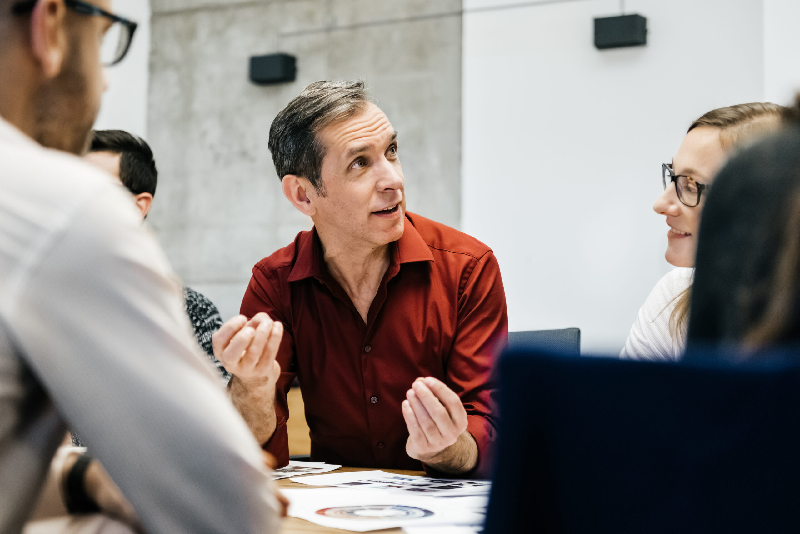 A mature man is speaking in a business meeting surrounded by colleagues listening to him. He uses his hands and seems convinced and focused.