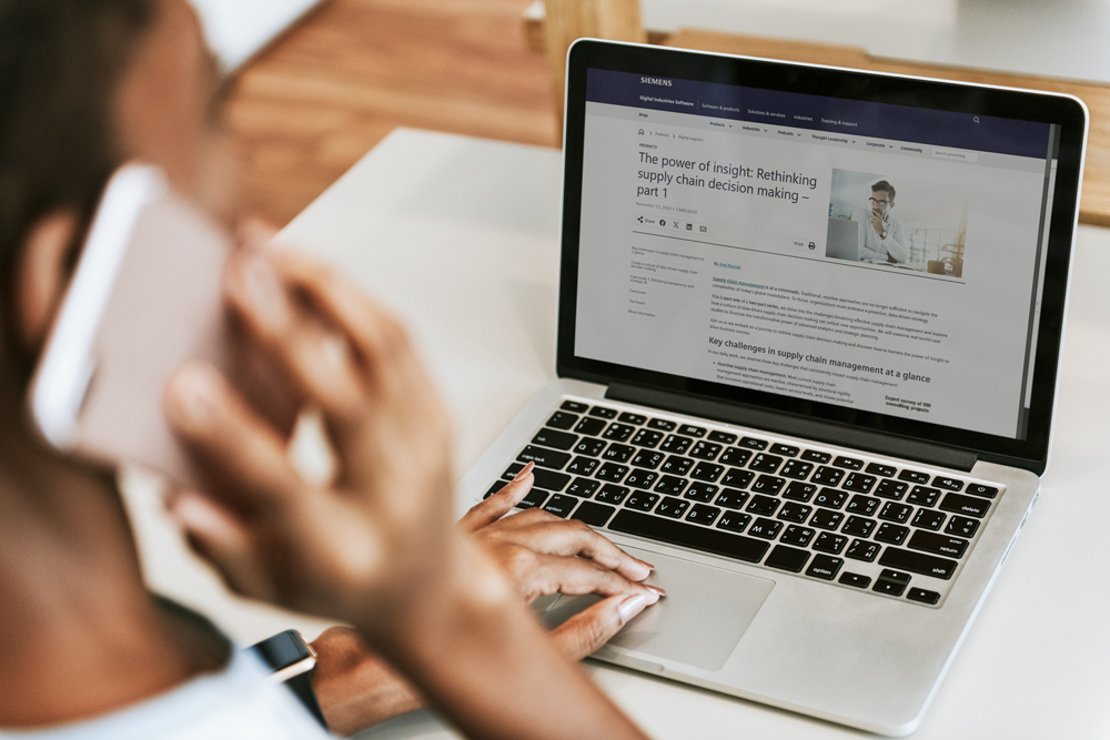 A person is using a laptop, reading an article titled "The power of insight: Rethinking supply chain decision making - part 1," while holding a phone to their ear. The background is slightly blurred, focusing on the laptop screen.
