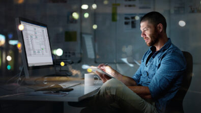 Man in blue denim shirt sitting at desk working on a model in Solid Edge 3D CAD software and holding a notepad